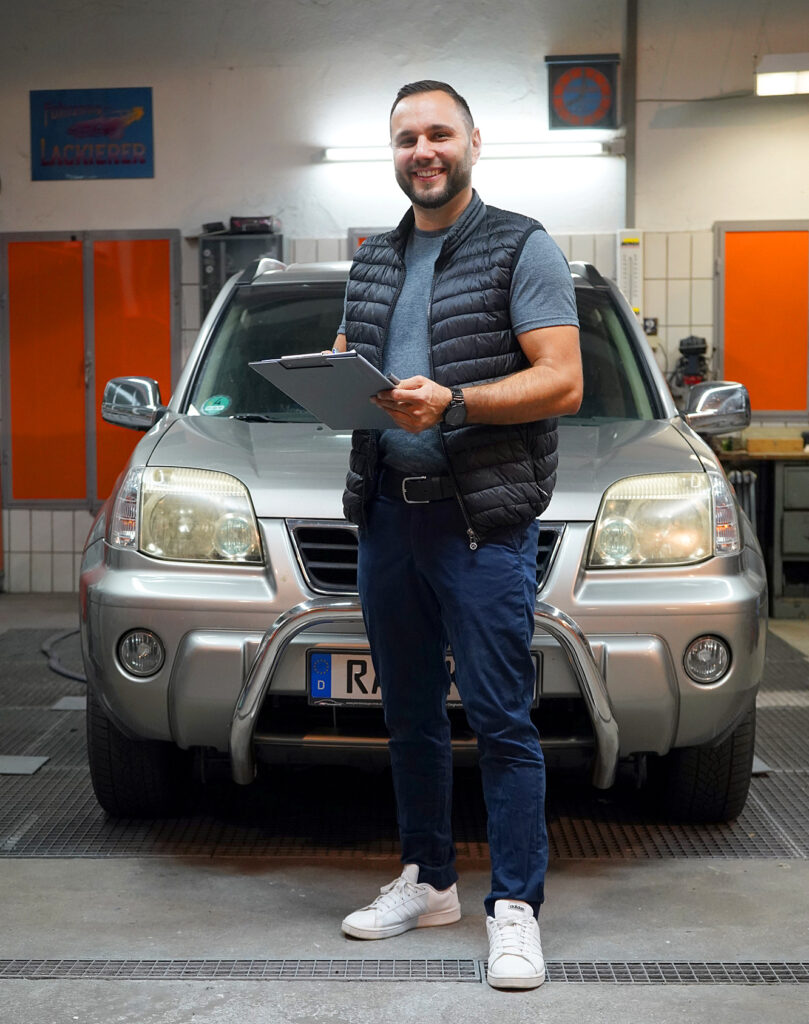 Kfz-Gutachter Baden steht vor einem Auto in der Werkstatt mit Schreibblock in der Hand